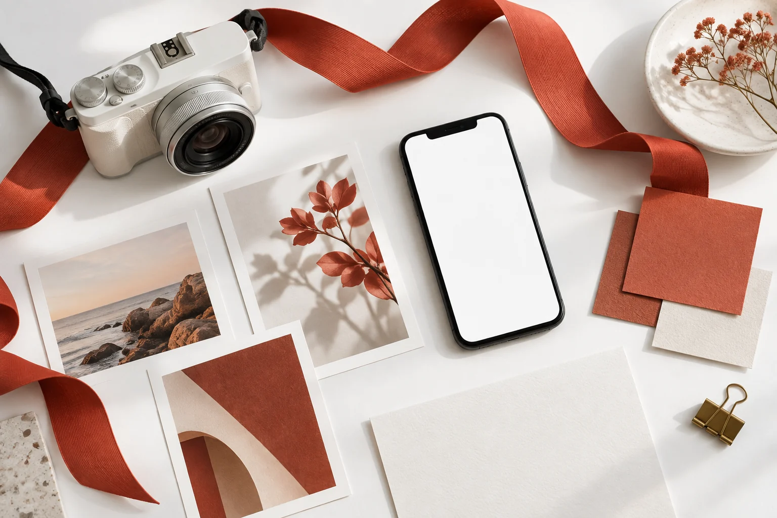 Illustrative white studio flat lay with a camera, blank smartphone, photographs, coral ribbon and tactile colour swatches.